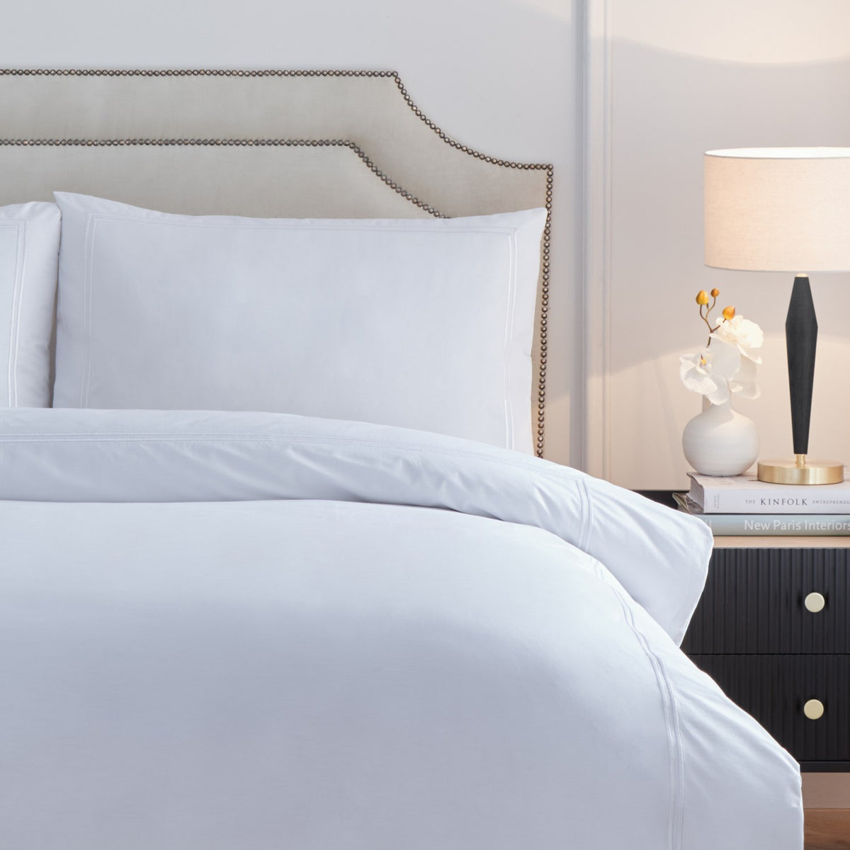 Elara white cotton duvet cover set with embroidered border detail on cover and pillowcases, styled on a bed beside a dark headboard and nightstand lamp.