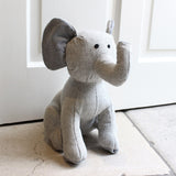 Elephant Animal Wool Door Stop in Grey, a sitting elephant with raised trunk and large ears in herringbone fabric, shown beside a white panelled door.
