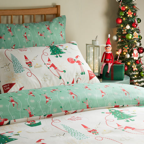 Elfie Printed Reversible Duvet Cover Set in Cream/Mint on a bed, cream side showing playful elf and North Pole scenes, mint reverse with elf print, styled beside a Christmas tree and elf toy.