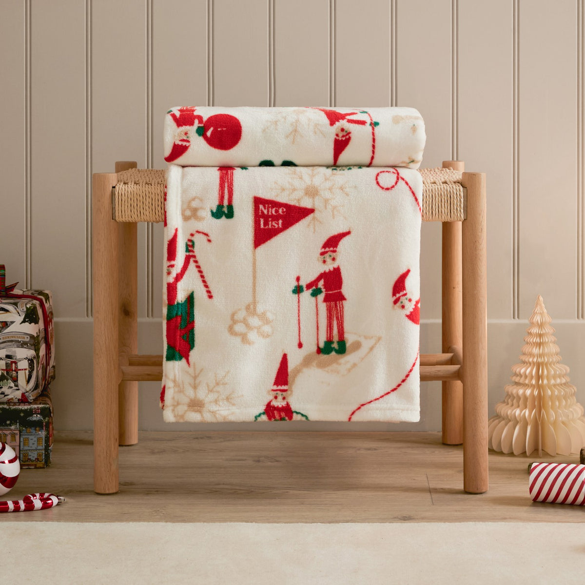 Elfie Printed Polyester Throw in cream, draped over a wooden bench with a festive red and green elf print, shown in a Christmas-themed room setting.