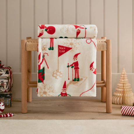 Elfie Printed Polyester Throw in cream, draped over a wooden bench with a festive red and green elf print, shown in a Christmas-themed room setting.