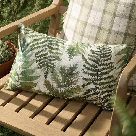Elowen Linen Cotton Cushion in Moss, a rectangular lumbar cushion with a hand-painted fern and foliage design in green tones, shown on a wooden garden bench beside a moss check cushion.