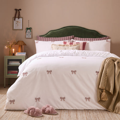 White cotton blend duvet cover set with embroidered burgundy and pink bows, styled on a bed with checked pillowcases in a warm-toned bedroom.