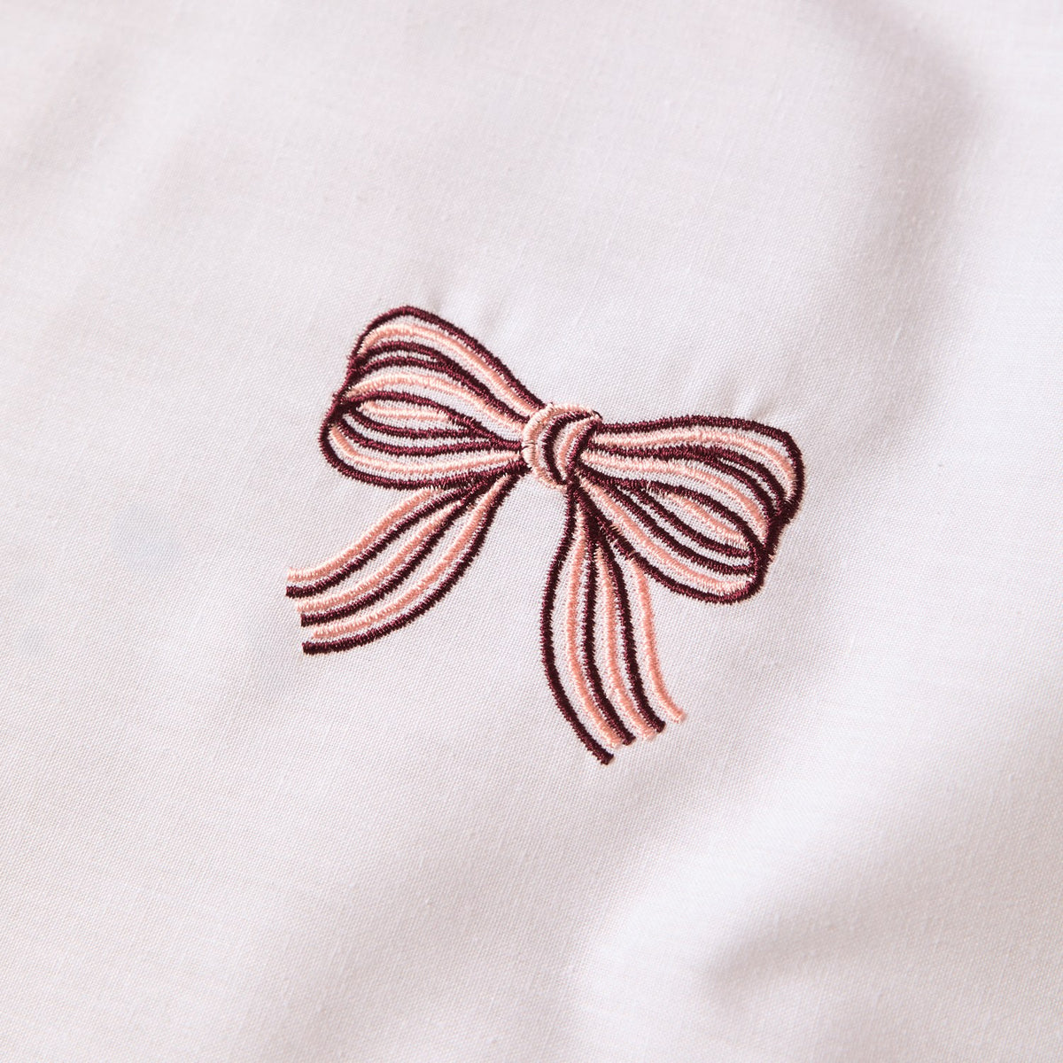 Close-up of the Embroidered Bows Cotton Blend Duvet Cover Set in Burgundy/Pink, showing a single ribbon bow embroidered in burgundy and pink thread on a white cotton blend ground.