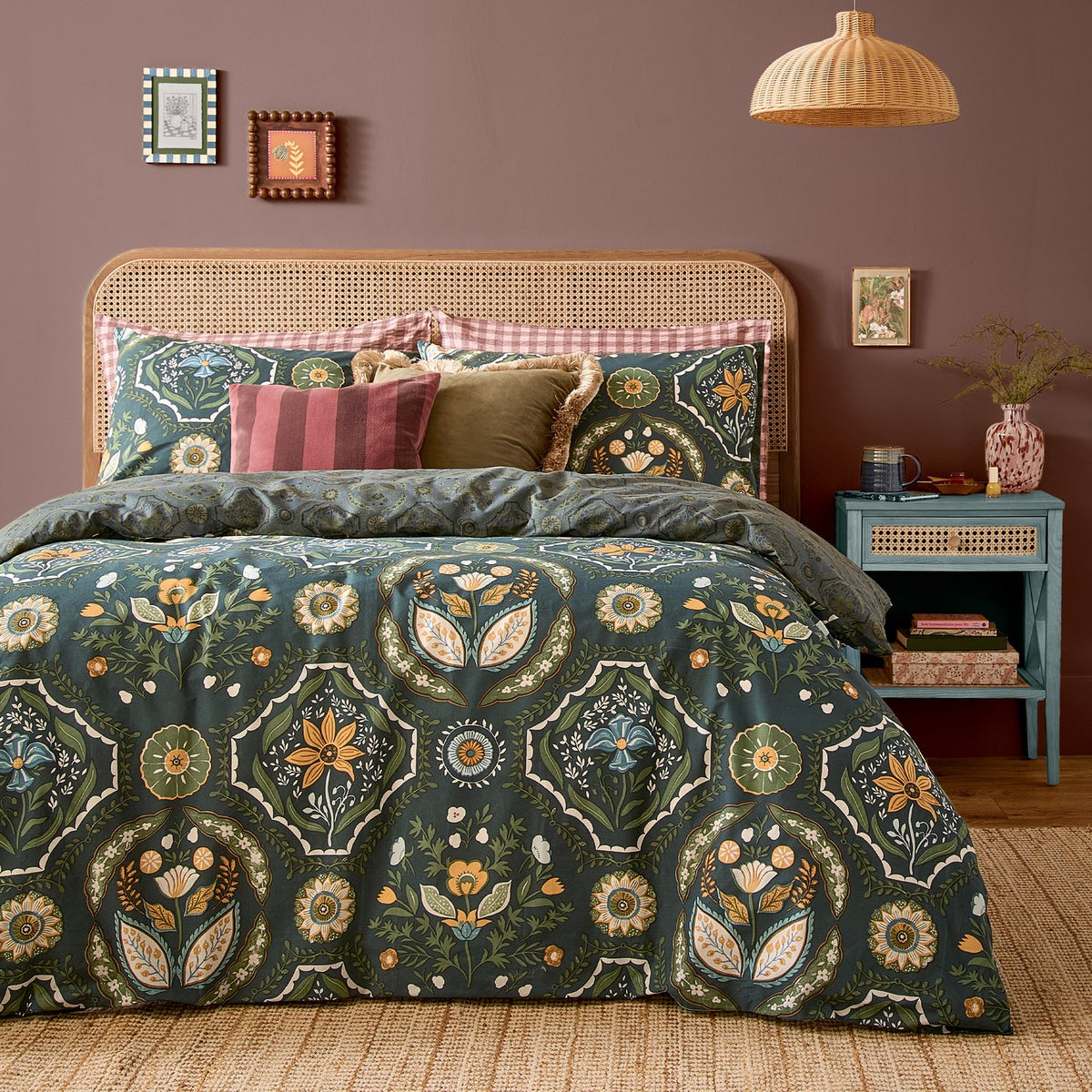 Ember Reversible Floral Duvet Cover Set in midnight green, with symmetrical amber, sage and blue floral panels, styled on a bed with a rattan headboard and pink gingham trim pillowcases.