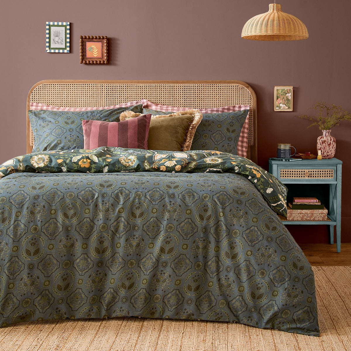 Ember Reversible Floral Duvet Cover Set in Midnight Green on a bed with matching pillowcases, styled with velvet cushions, rattan headboard and teal bedside table.