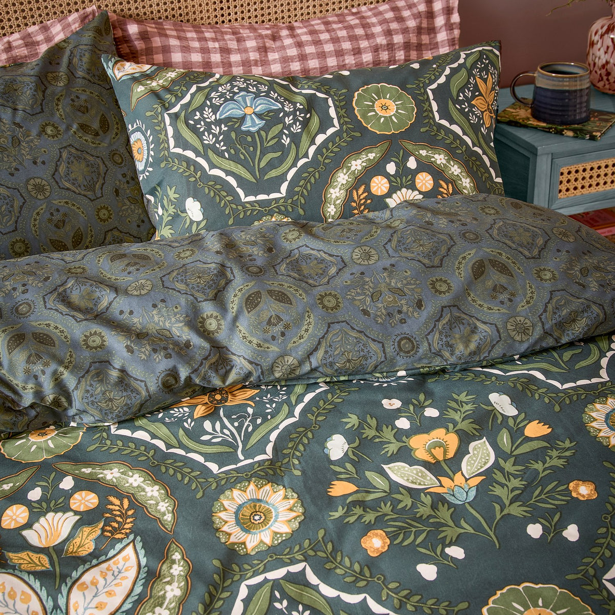 Ember Reversible Floral Duvet Cover Set in midnight green, styled on a bed with matching pillowcases showing tulip and rosette panels in amber, sage, blue and cream.