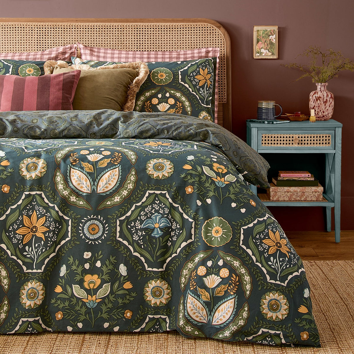 Ember Reversible Floral Duvet Cover Set in Midnight Green with matching pillowcases, featuring symmetrical amber, sage, blue and cream floral panels, styled on a bed with a rattan headboard.