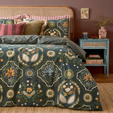 Ember Reversible Floral Duvet Cover Set in Midnight Green with matching pillowcases, featuring symmetrical amber, sage, blue and cream floral panels, styled on a bed with a rattan headboard.