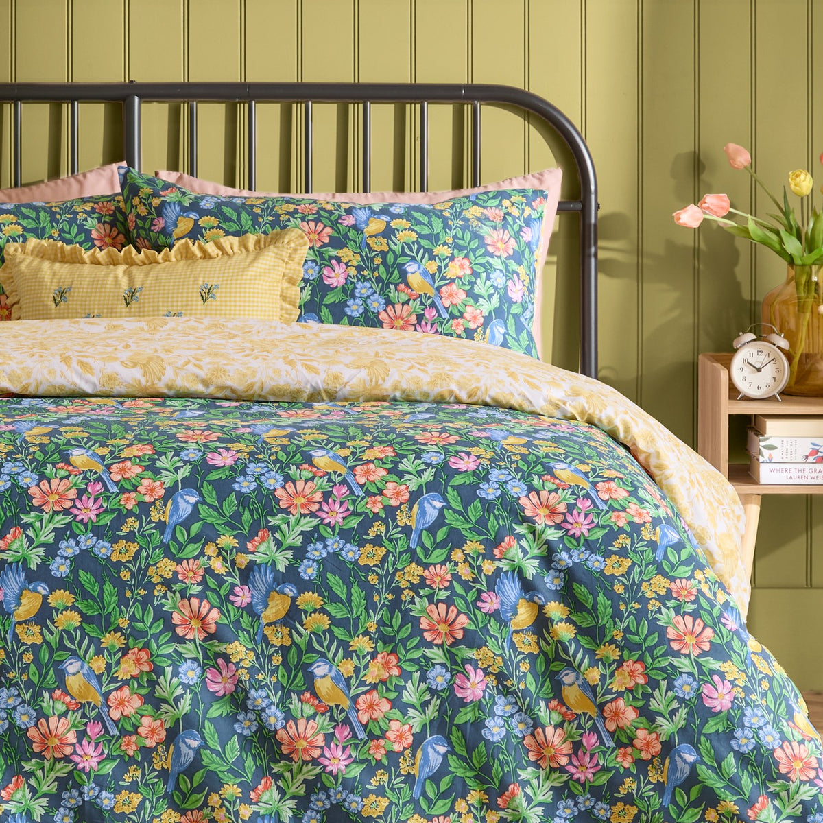 Emerie Reversible Floral Duvet Cover Set in navy, shown on a bed with pillowcases in a bedroom, featuring pink, coral and yellow blooms with birds among green foliage on a dark ground.