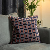 Empire Jacquard Velvet Cushion in blush and navy, square with a geometric octagon and square pattern, shown on a grey sofa beside a curtain and potted plant.