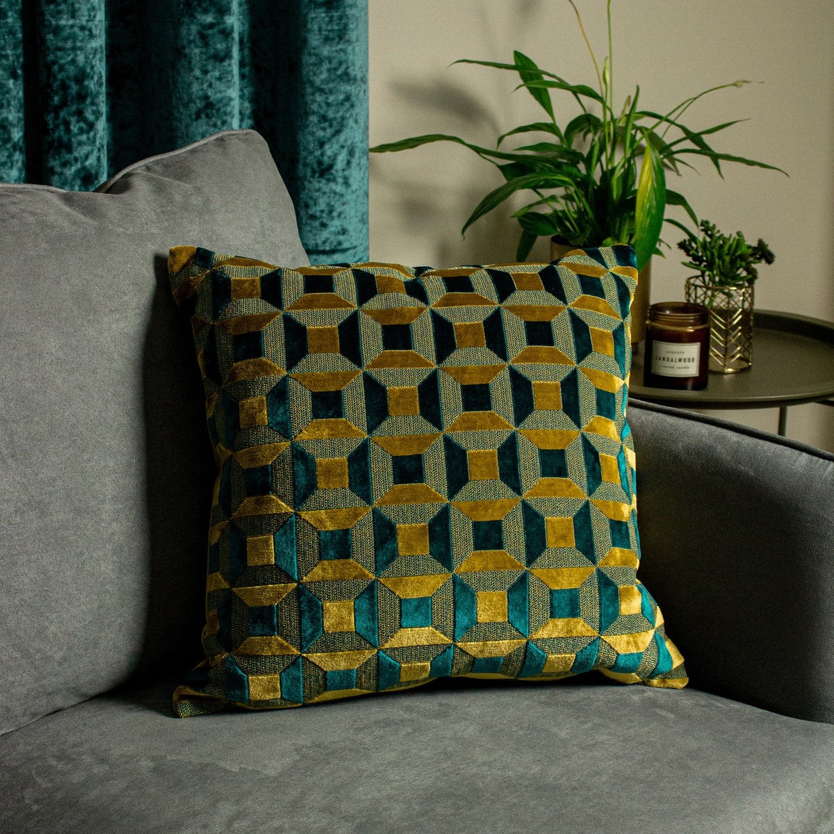 Empire Jacquard Velvet Cushion in teal and gold, square with geometric block pattern, shown on a grey sofa beside a teal curtain and potted plant.