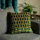 Empire Jacquard Velvet Cushion in teal and gold, square with geometric block pattern, shown on a grey sofa beside a teal curtain and potted plant.