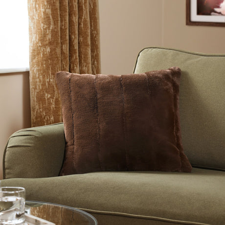 Empress Plain Faux Fur Cushion in brown, square with dense textured pile, shown on an olive green sofa in a living room setting.