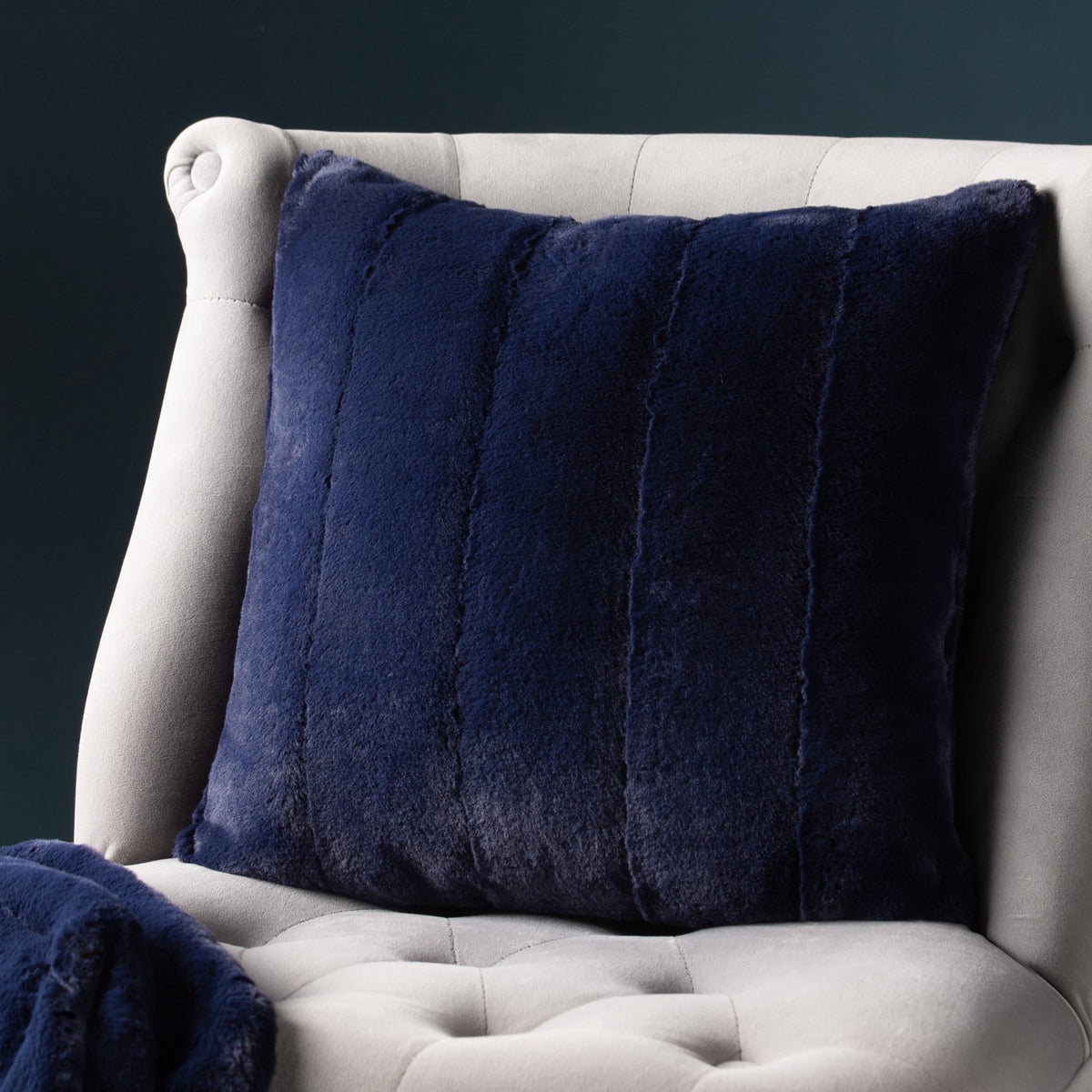 Empress Velvet Faux Fur Cushion in navy, square with vertical channel stitching, shown on a light grey upholstered armchair.