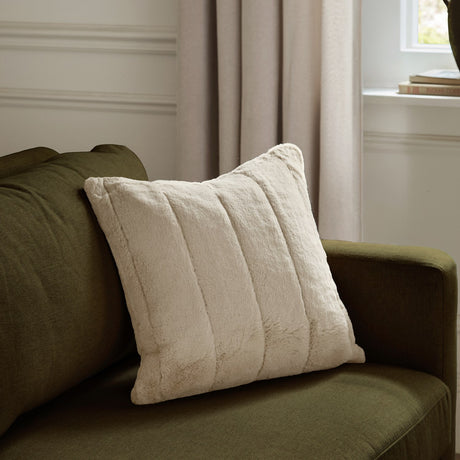 Empress Plain Faux Fur Cushion in oatmeal with ribbed vertical channels, shown on a green sofa beside a curtained window.