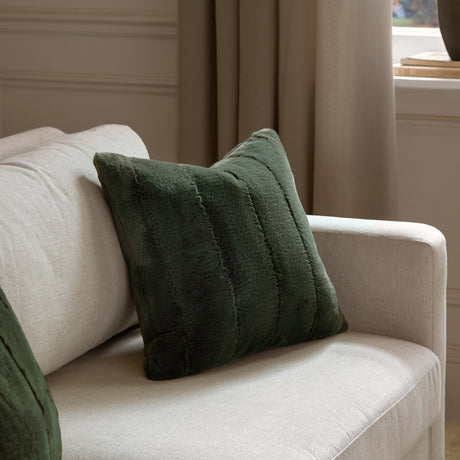Empress Plain Faux Fur Cushion in pine green, square with ridged vertical channels, styled on a beige sofa in a living room setting.