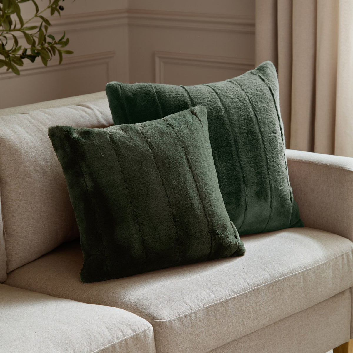 Two Empress square faux fur cushions in pine green with vertical panelled texture, styled on a beige fabric sofa in a living room.