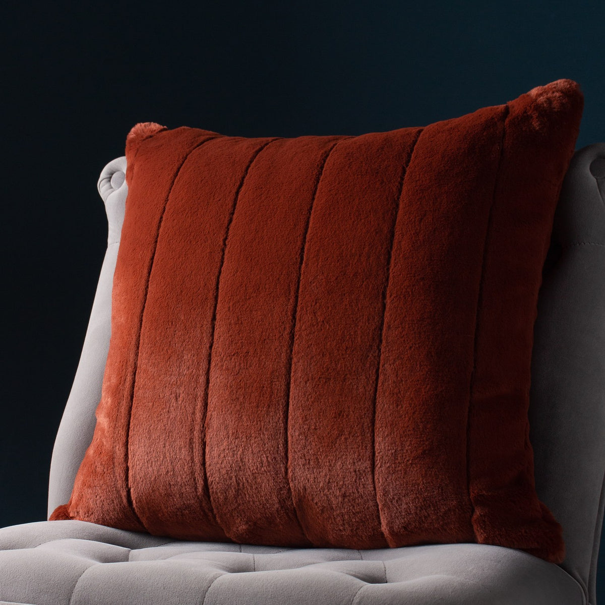 Empress Velvet Faux Fur Cushion in rust orange with vertical ribbed channels, shown on a light grey upholstered chair against a dark teal background.