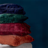 Empress Velvet Faux Fur Cushion in rust orange, square with ribbed vertical channels, shown stacked with navy, green, and burgundy cushions against a dark teal background.