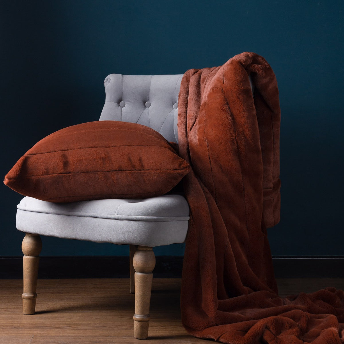 Empress Velvet Faux Fur Cushion in rust with ribbed channel front, styled on a grey armchair with a matching faux fur throw against a dark teal wall.