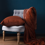 Empress Velvet Faux Fur Cushion in rust with ribbed channel front, styled on a grey armchair with a matching faux fur throw against a dark teal wall.