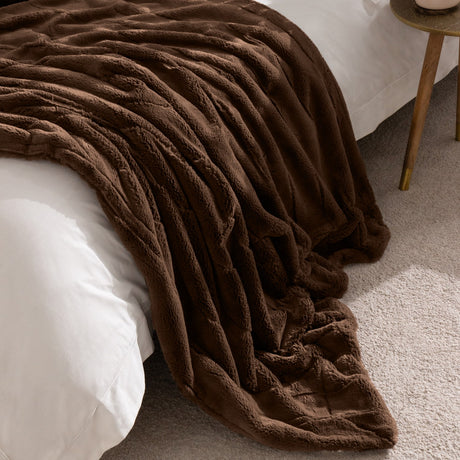 Empress Plain Faux Fur Throw in brown, dense pile with soft vertical panels, draped over a white bed beside a wooden side table.