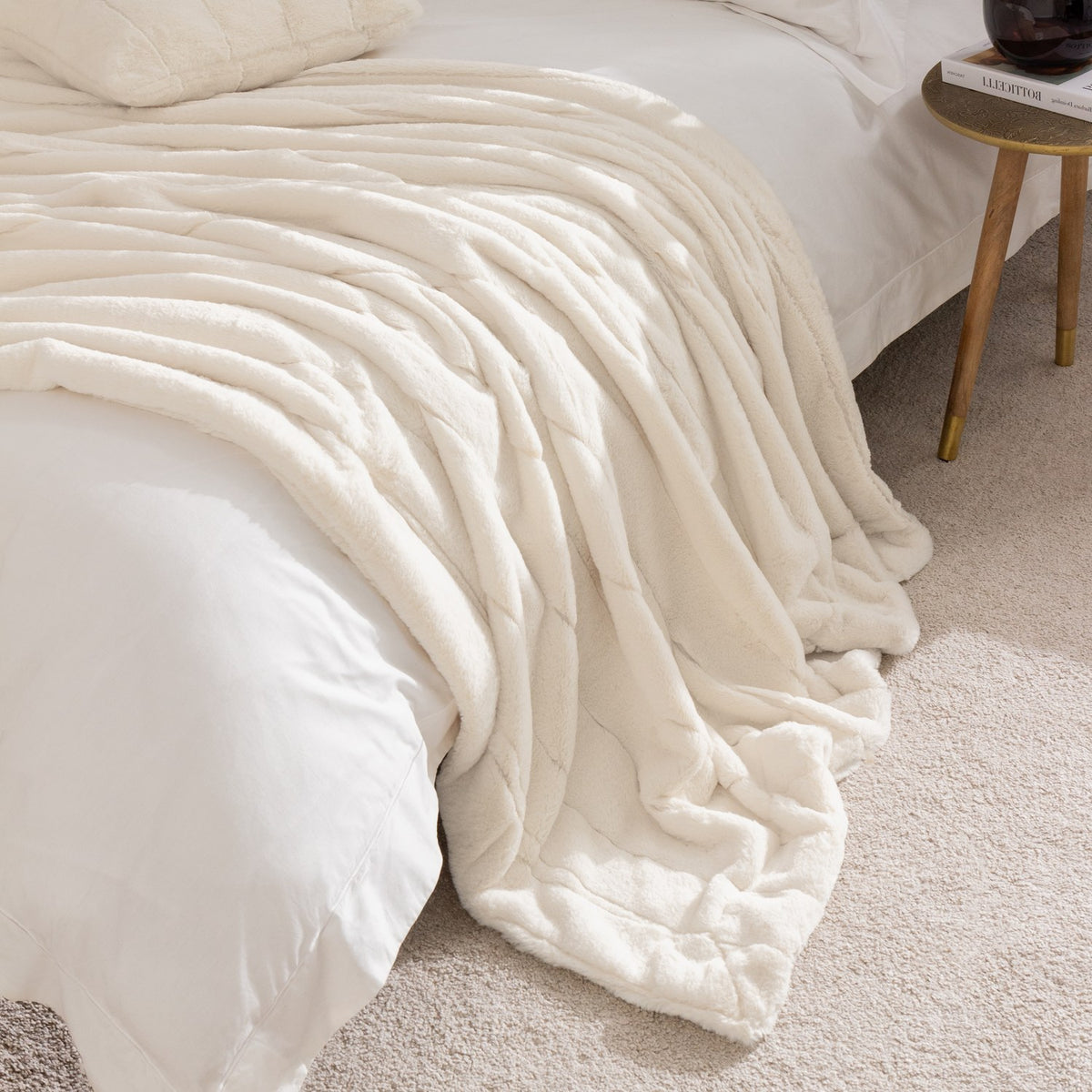 Empress Velvet Faux Fur Throw in cream, draped over a bed with ribbed, sculpted channels, shown in a bedroom beside a wooden side table.