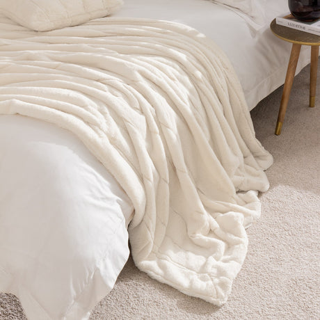 Empress Velvet Faux Fur Throw in cream, draped over a bed with ribbed, sculpted channels, shown in a bedroom beside a wooden side table.