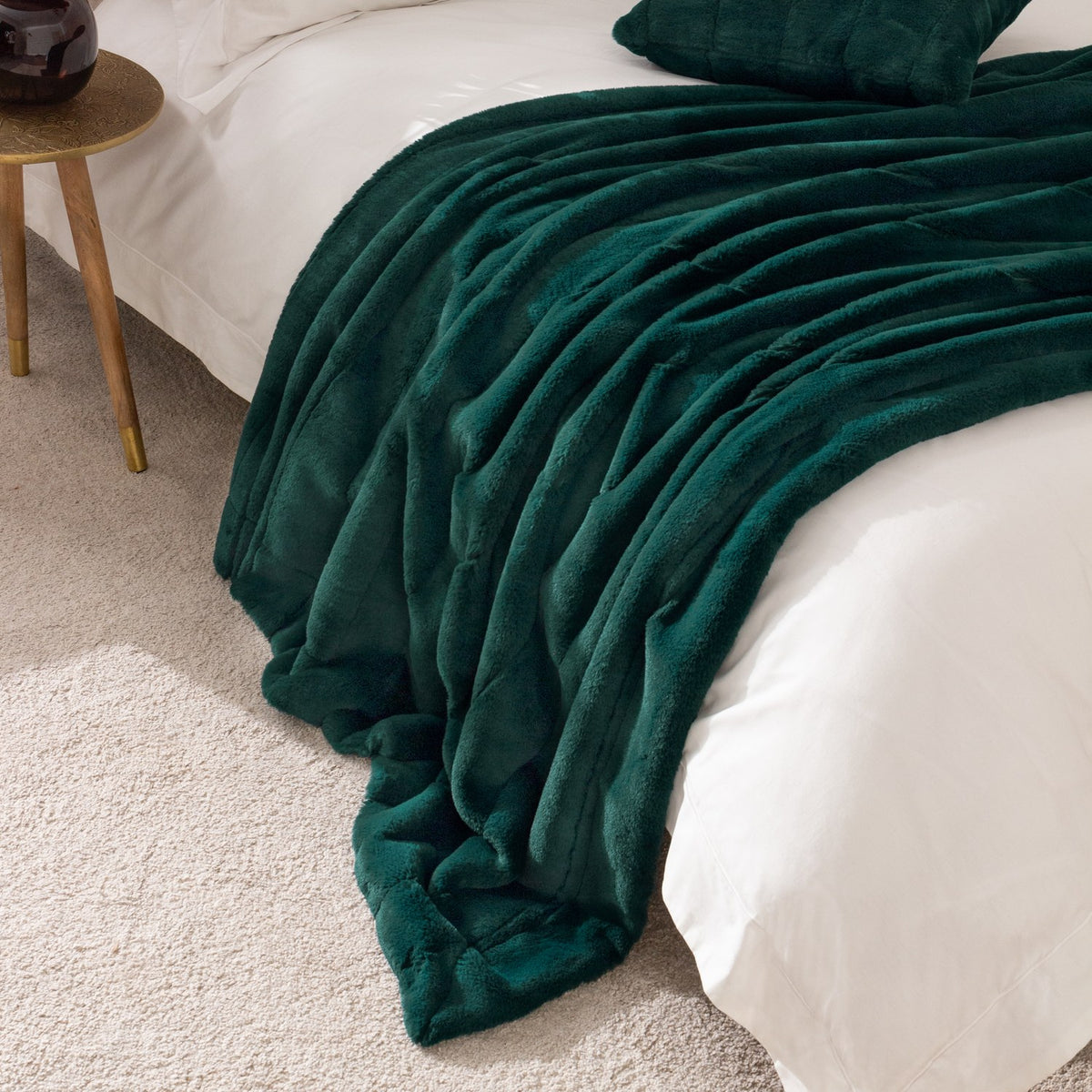Empress Velvet Faux Fur Throw in emerald green draped over a white bed with matching cushion, faux fur pile texture visible, styled beside a wooden side table.