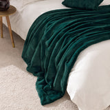 Empress Velvet Faux Fur Throw in emerald green draped over a white bed with matching cushion, faux fur pile texture visible, styled beside a wooden side table.