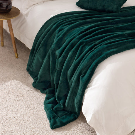 Empress Velvet Faux Fur Throw in emerald green draped over a white bed with matching cushion, faux fur pile texture visible, styled beside a wooden side table.