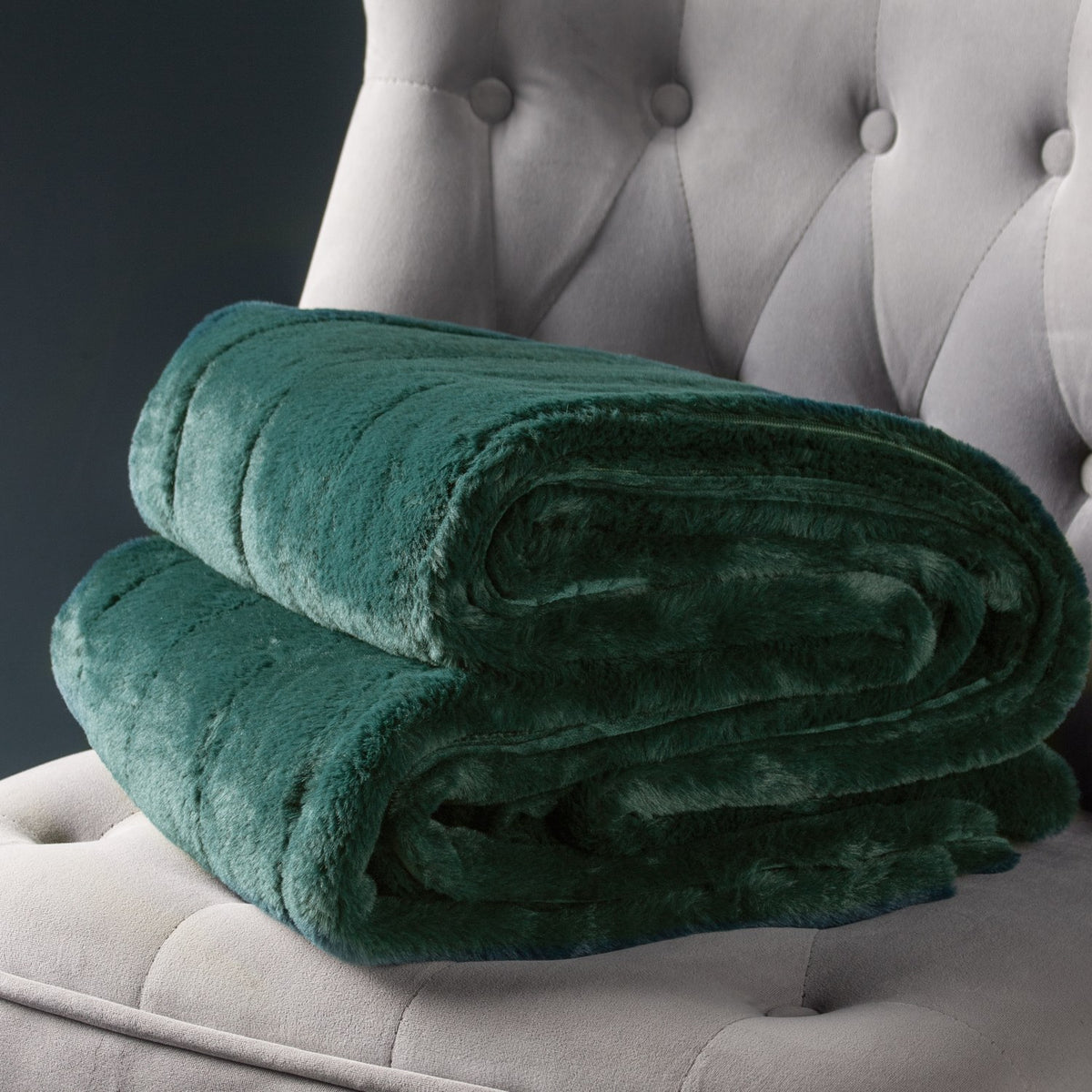 Empress Velvet Faux Fur Throw in emerald green, folded to show its soft pile texture, displayed on a light grey tufted armchair.