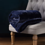 Empress Velvet Faux Fur Throw in navy, folded on a grey tufted armchair, showing its plush textured pile and quilted stripe pattern.