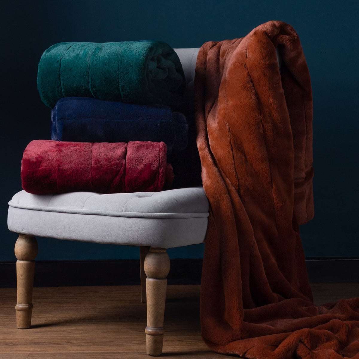 Empress Velvet Faux Fur Throw in navy, rolled and stacked with green and red versions on a light upholstered stool, beside a draped rust-coloured throw against a dark blue wall.