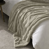 Empress Plain Faux Fur Throw in oatmeal, a ribbed dense-pile blanket draped over a white bedspread in a bedroom setting with a wooden side table nearby.