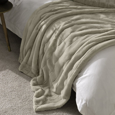 Empress Plain Faux Fur Throw in oatmeal, a ribbed dense-pile blanket draped over a white bedspread in a bedroom setting with a wooden side table nearby.