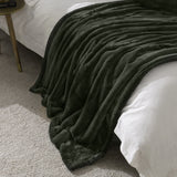 Empress Plain Faux Fur Throw in pine green, dense plush pile draped over a bed corner with white bedding, gold side table visible in a bedroom setting.
