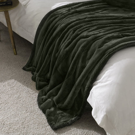 Empress Plain Faux Fur Throw in pine green, dense plush pile draped over a bed corner with white bedding, gold side table visible in a bedroom setting.