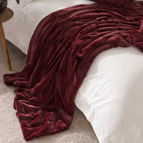 Empress Velvet Faux Fur Throw in ruby red draped over a white bed, showing ribbed plush pile texture and folded fabric edge, styled in a bedroom setting.