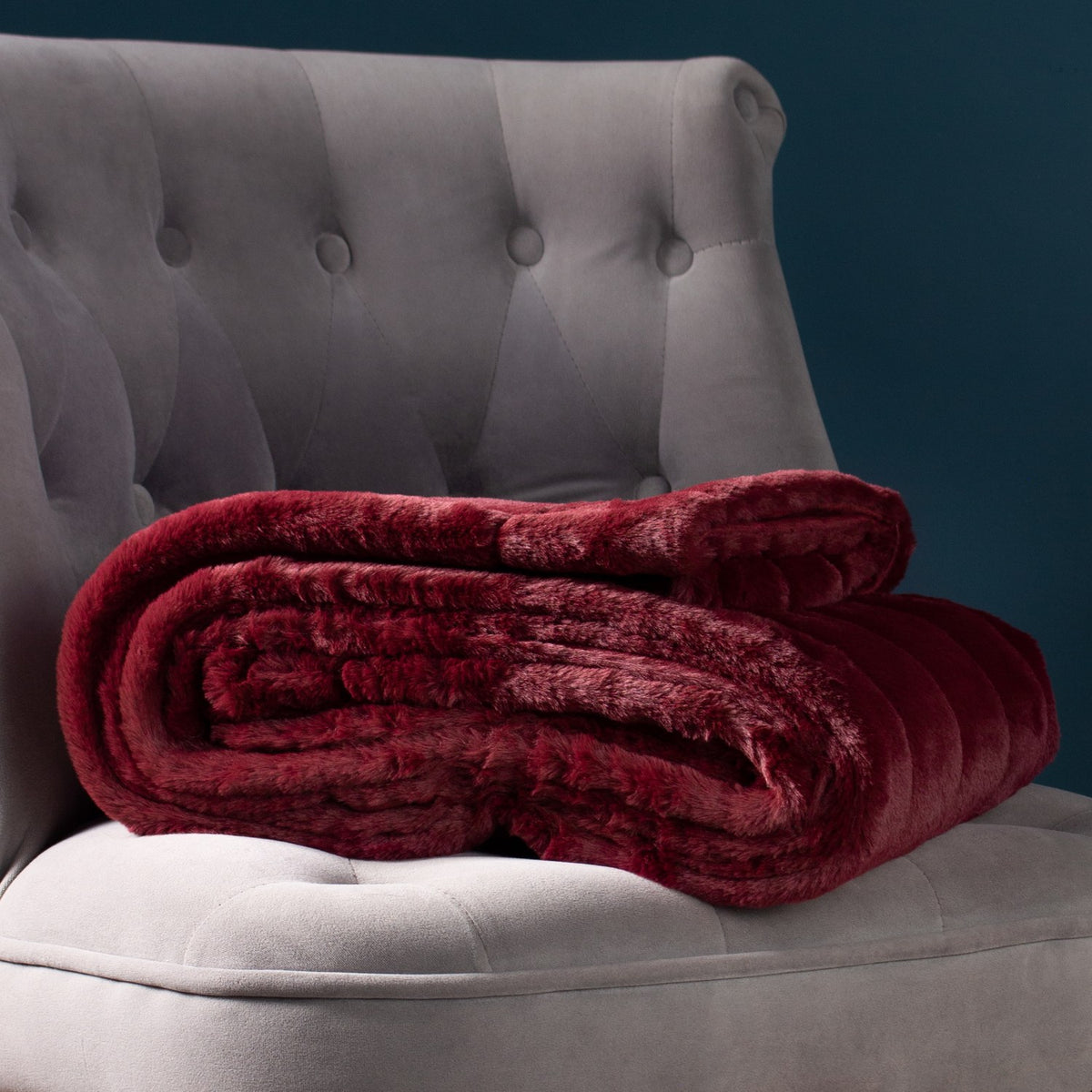 Empress Velvet Faux Fur Throw in ruby red, folded with plush textured pile, shown on a grey tufted armchair against a dark teal background.