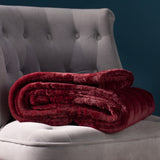 Empress Velvet Faux Fur Throw in ruby red, folded with plush textured pile, shown on a grey tufted armchair against a dark teal background.