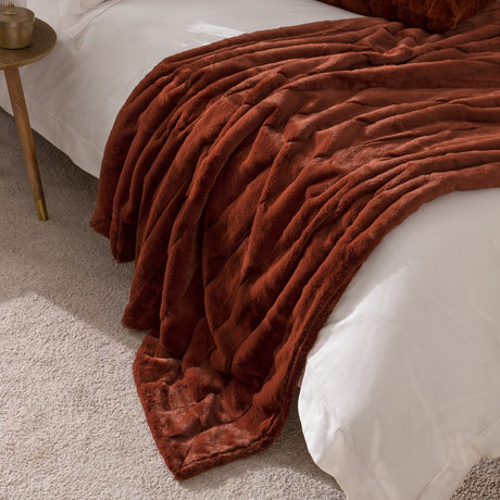 Empress Velvet Faux Fur Throw in rust draped over a white bed, showing ribbed channel texture on the pile in a bedroom setting.
