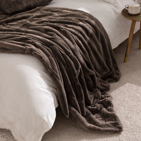 Empress Velvet Faux Fur Throw in taupe brown, channelled deep-pile faux fur texture, draped over a white bed beside a wooden side table in a bedroom setting.