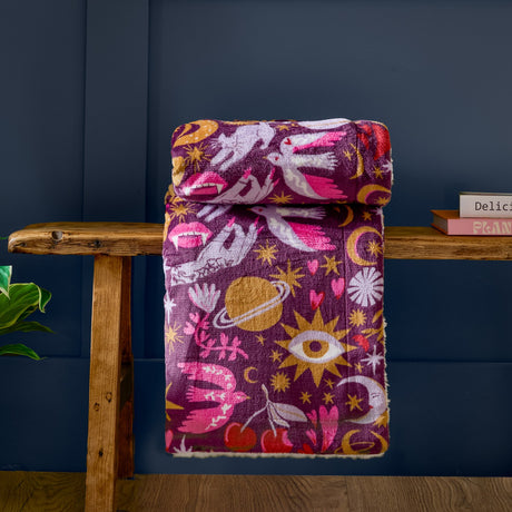 Enchanted Printed Polyester Throw in pink, folded on a wooden bench with a mystical print of eyes, moons, doves and stars on a deep plum ground.