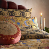 Ethereal Reversible Abstract Duvet Cover Set in Gold Rust, styled on a bed with celestial sun and moon print pillowcases, a round sun-embroidered cushion, and lit candles beside it.