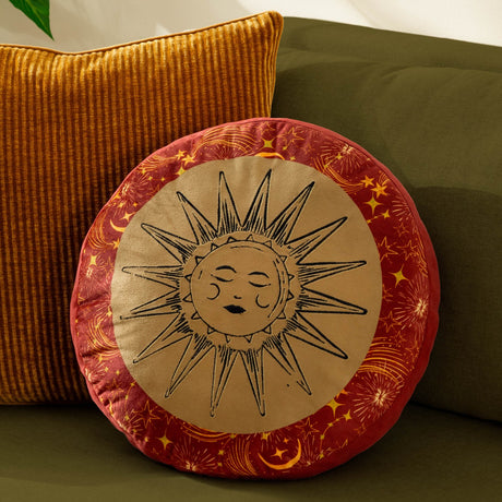 Round velvet cushion in gold and rust from the Ethereal range, featuring an embroidered sun face on a gold panel edged with a star and moon print border, shown on a green sofa.