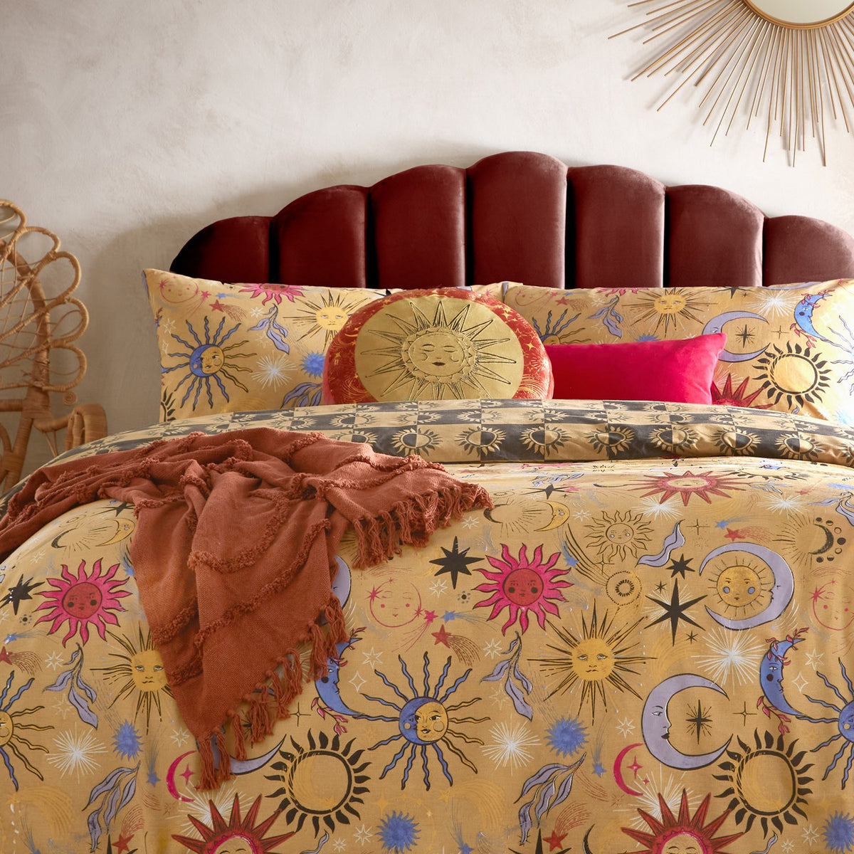 Round Ethereal Abstract Velvet Cushion in gold and rust, featuring an embroidered sun face on a gold panel with a rust border of moons and stars, styled on a celestial-print bed.