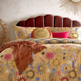 Round Ethereal Abstract Velvet Cushion in gold and rust, featuring an embroidered sun face on a gold panel with a rust border of moons and stars, styled on a celestial-print bed.