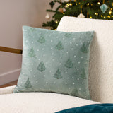 Square velvet cushion in mint green with embroidered fir trees and scattered pale dots, shown on a cream sherpa armchair beside a lit Christmas tree.